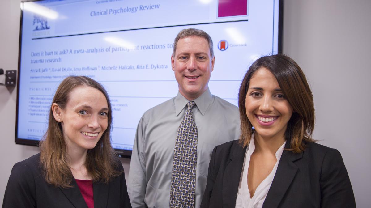 Anna Jaffe, David DiLillo, and Michelle Haikalis stand in front of a board displaying a journal publication. All three are smiling and wearing business attire.