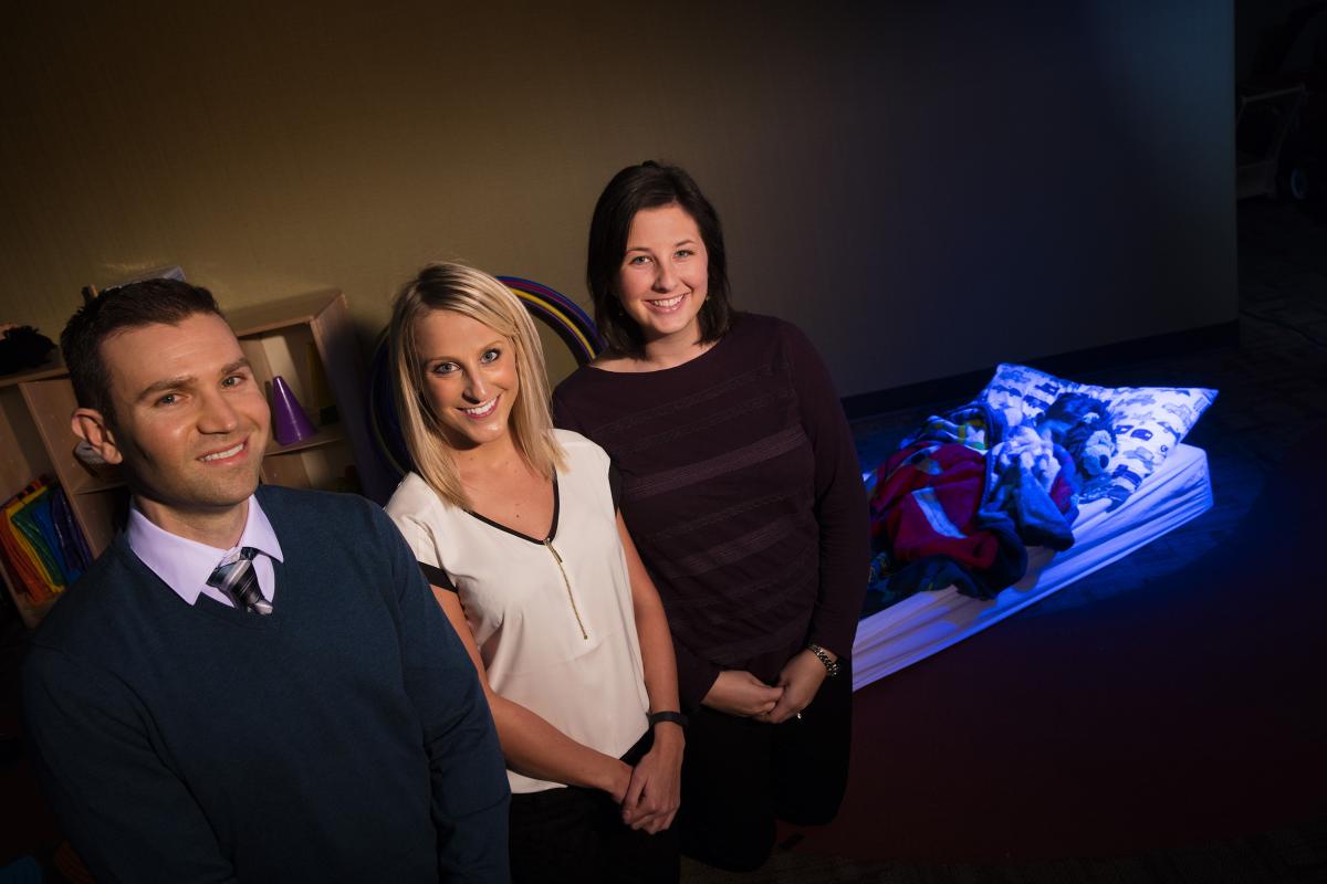 Tim Nelson, Alyssa Lundahl, and Katie Kidwell stand in front of a child's bed. The bed is illuminated by blue light and the people are lit by daylight. All three are smiling and wearing business casual attire.
