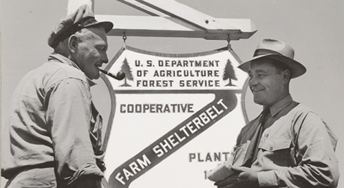 Black and white photo of two men in front of U.S. Department of Agriculture Forest Service sign