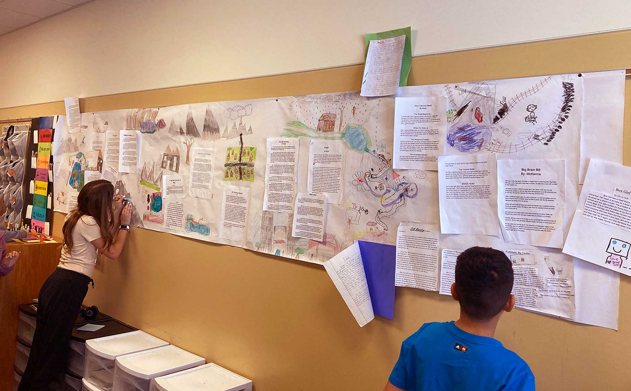 Two students stand at a wall filled with papers of sketch drawings and text. 