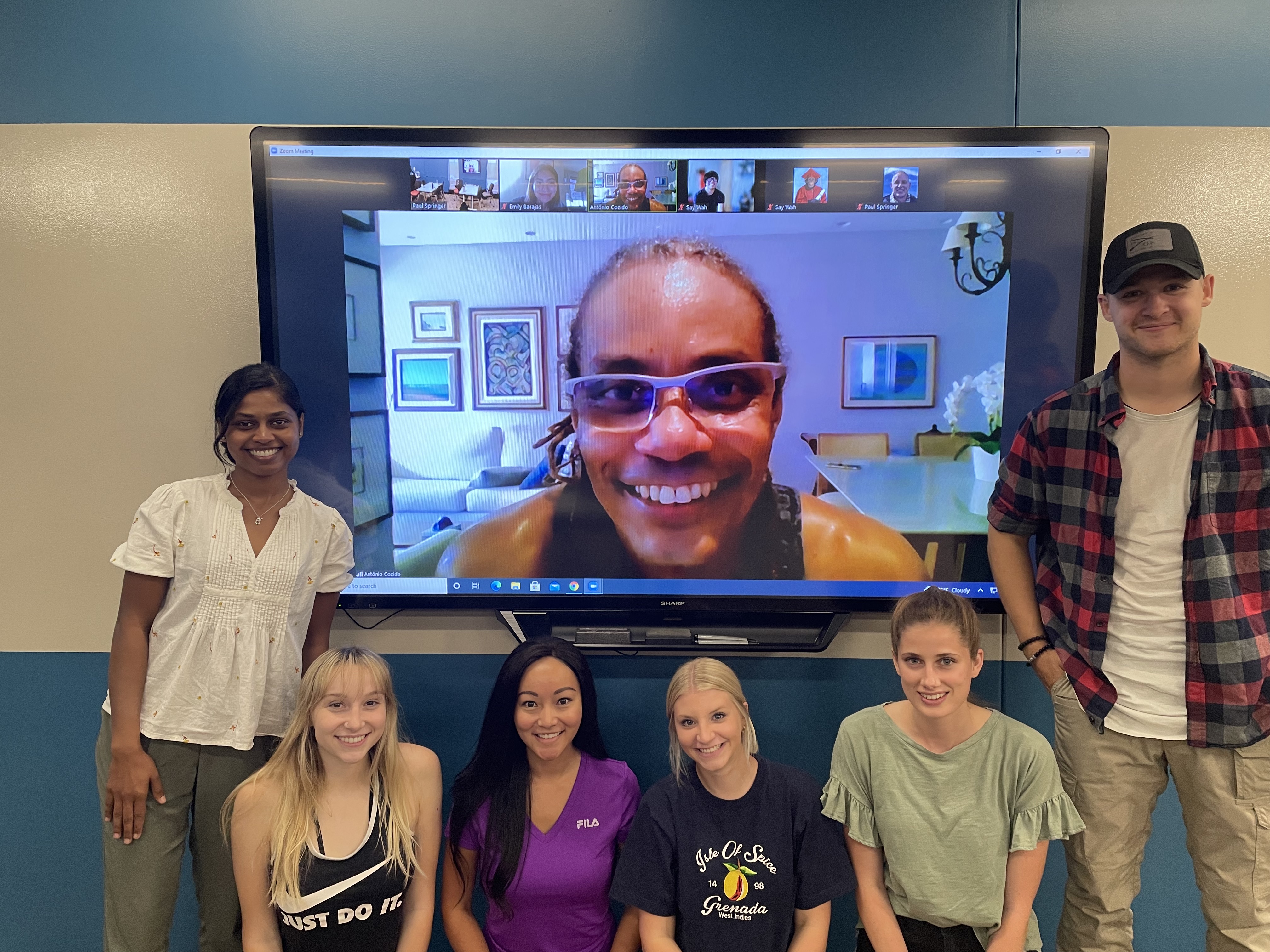 Six individuals standing in front of a blue and gray wall with a tv hanging on it. An additional person is on the tv screen smiling.