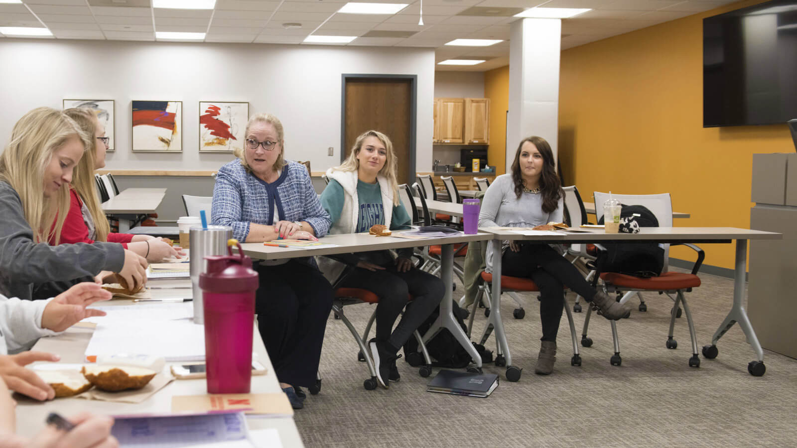 Sheree Moser sits at a table and speaks to students in a classroom featuring a mustard yellow wall. 