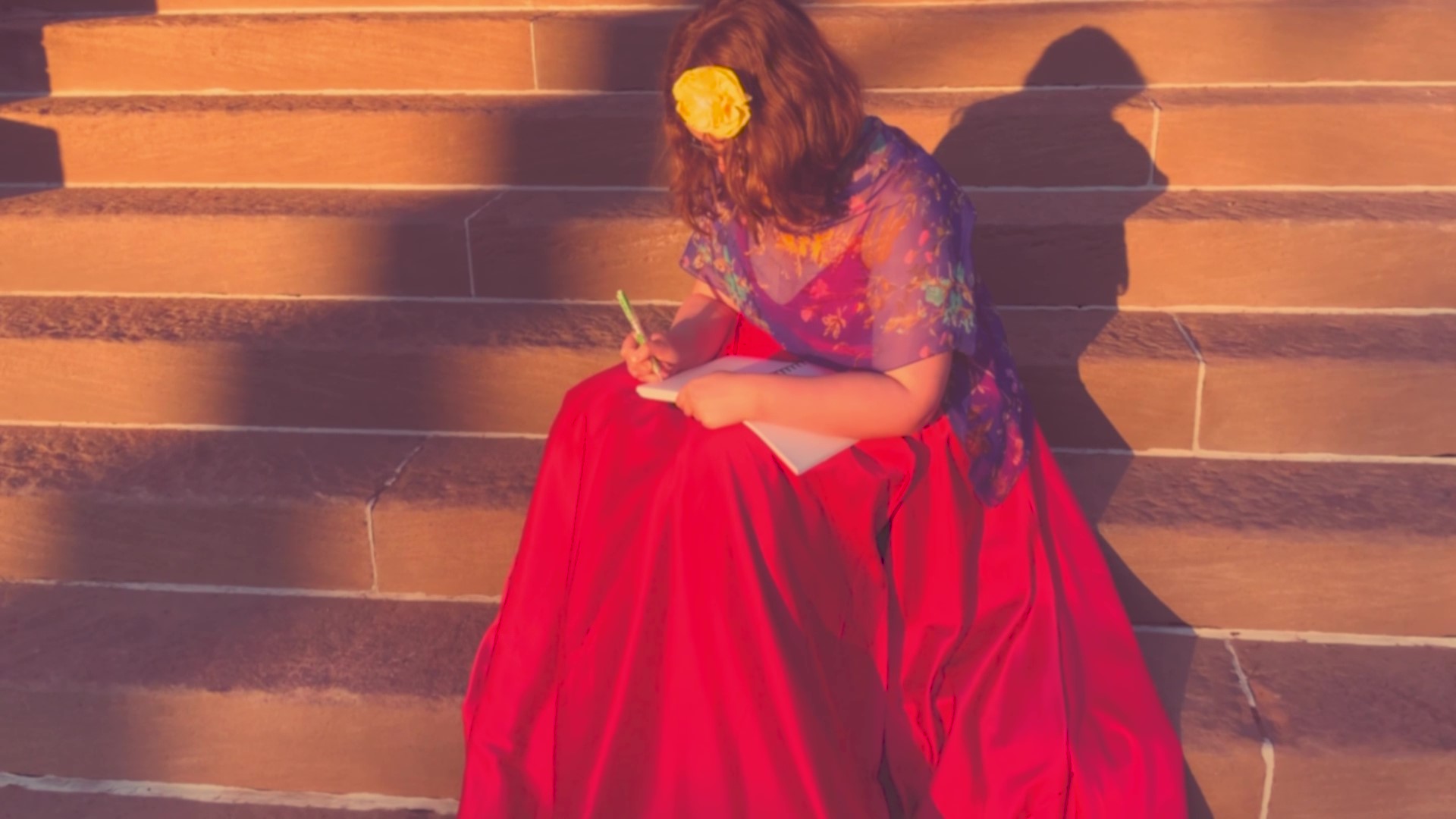 A person in a red dress sits writing on a flight of stairs outdoors. There is a yellow flower in their hair.