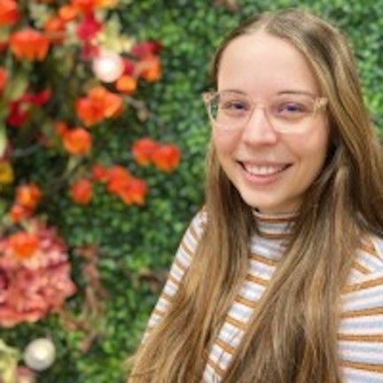 Kelly is pictured from the chest up smiling at the camera. She has long blonde hair and is wearing glasses. There is greenery and red flowers behind her in the background. 