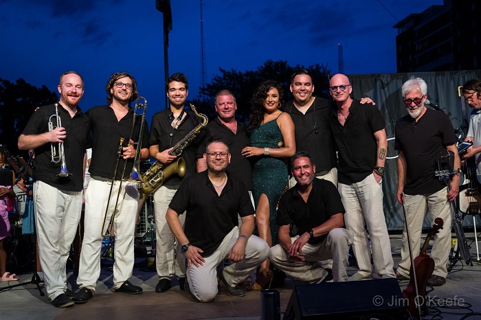 Alexis stands in the center of the photo in a green sparkly dress surrounded by her band dressed in black shirts and white pants on an outdoor stage with a darkening sky in the background